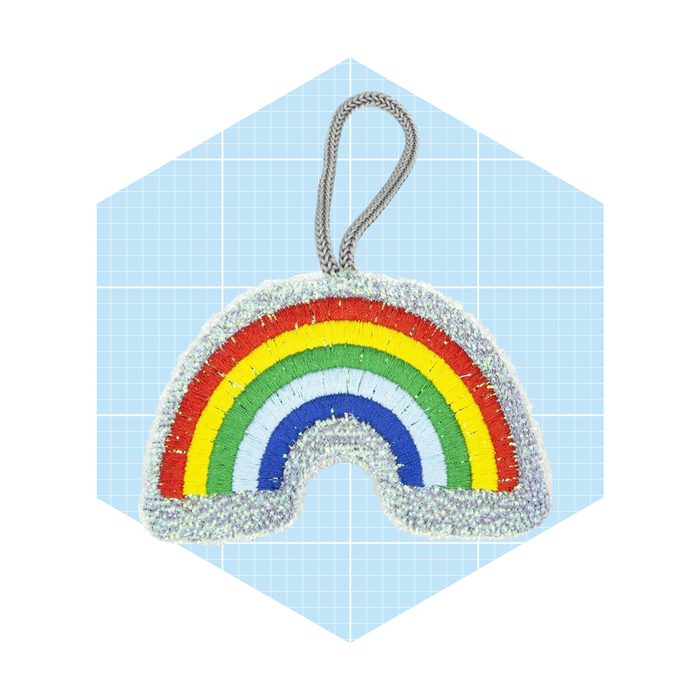 A colorful fabric rainbow-shaped ornament hangs against a light blue grid background, showcasing vibrant stripes of red, yellow, green, blue, and purple.