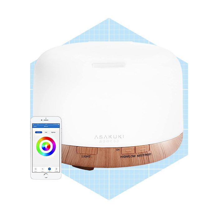 A white diffuser with wooden accents releases mist, while a smartphone displays color options nearby against a light blue grid background.