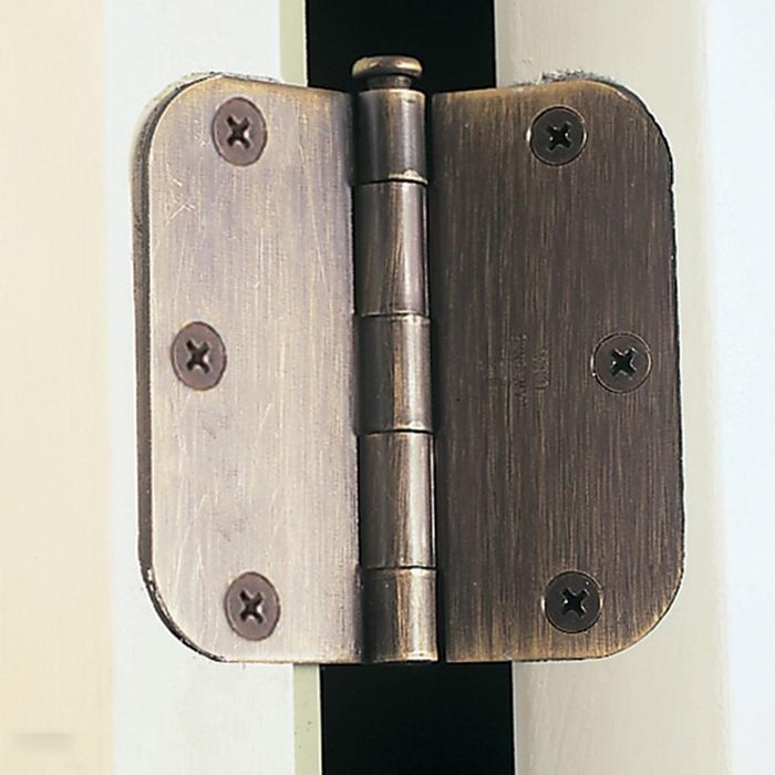 A close-up of a metal door hinge attached to the edge of a white door. The hinge is held in place by four screws, two on each side, with a brushed bronze finish. The door is slightly ajar, showing the hinge's full structure.